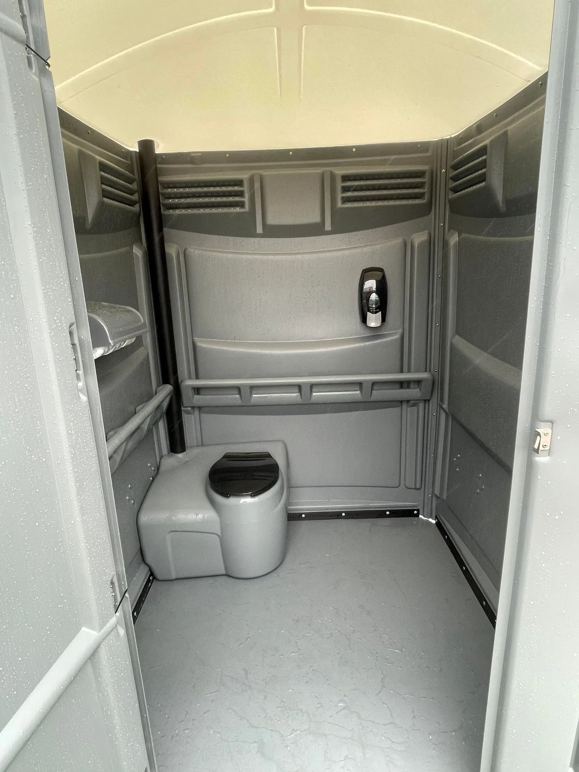 Inside view of a grey portable restroom with a single toilet, a soap dispenser on the wall, and a shelf.
