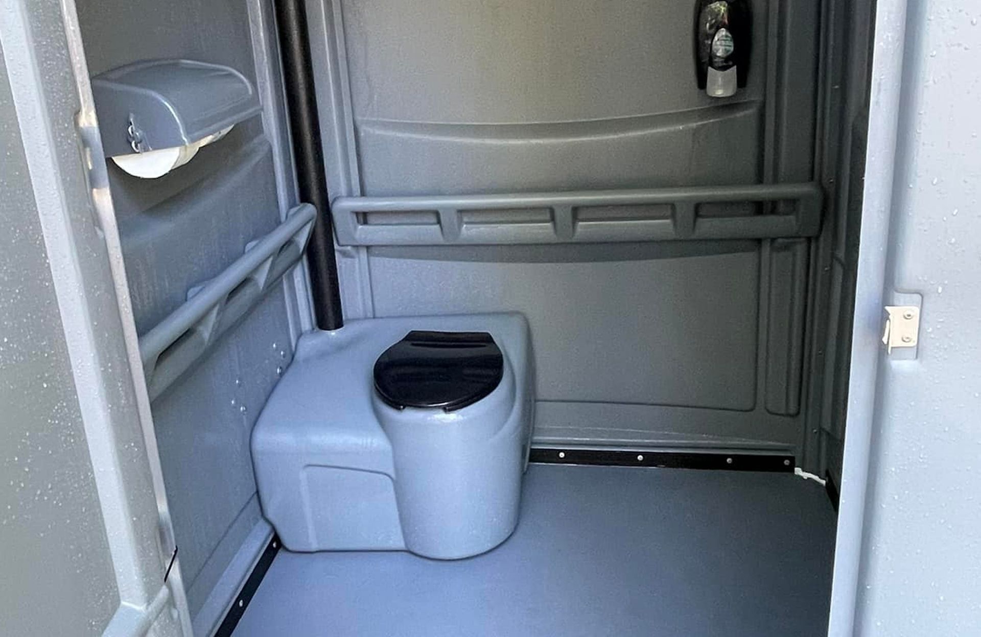 Interior of an accessible portable toilet with grab bars, a toilet seat, and a hand sanitizer dispenser on gray walls.