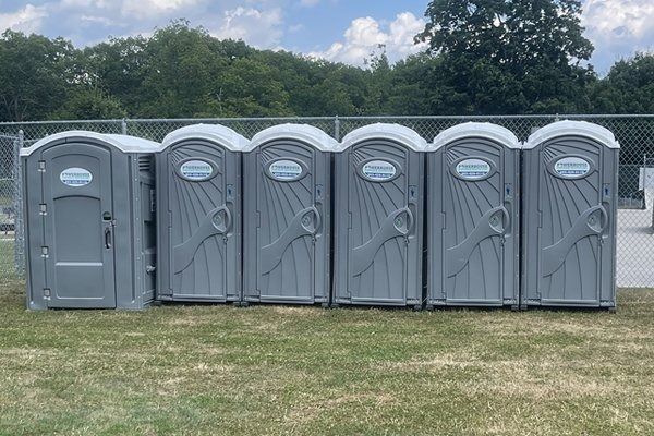 A row of six gray portable toilets lined up on grass in front of a chain-link fence under a cloudy blue sky.