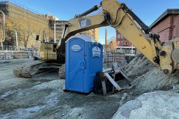 A blue portable toilet sits on a dirt construction site next to a large yellow CAT excavator under a clear blue sky.