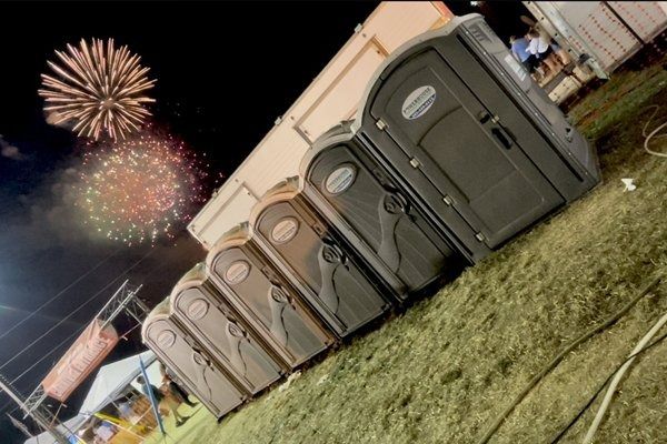 A row of portable toilets stands on grass at night, with bright fireworks exploding in the dark sky above.