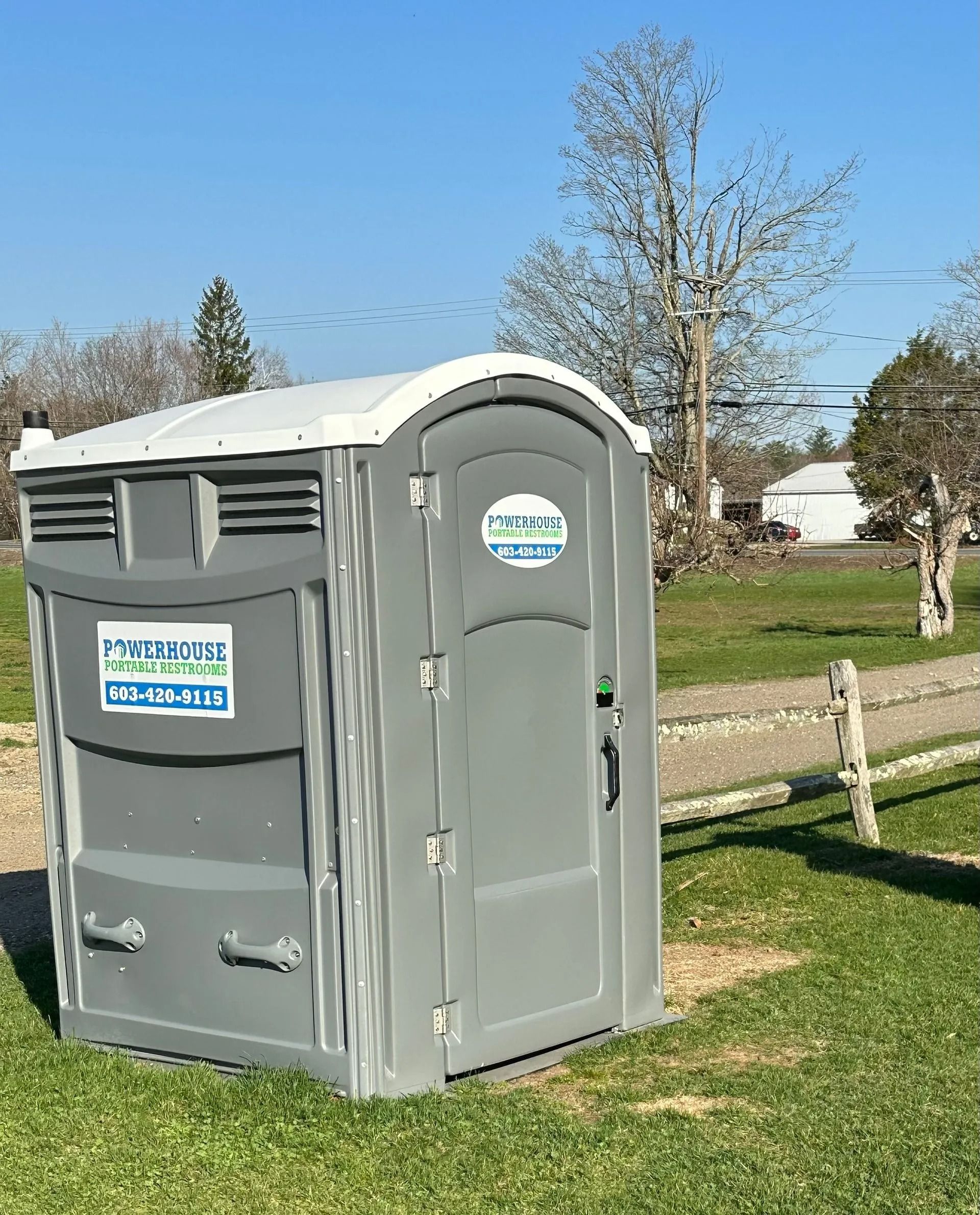 A gray portable toilet stands on a grassy field on a sunny day.