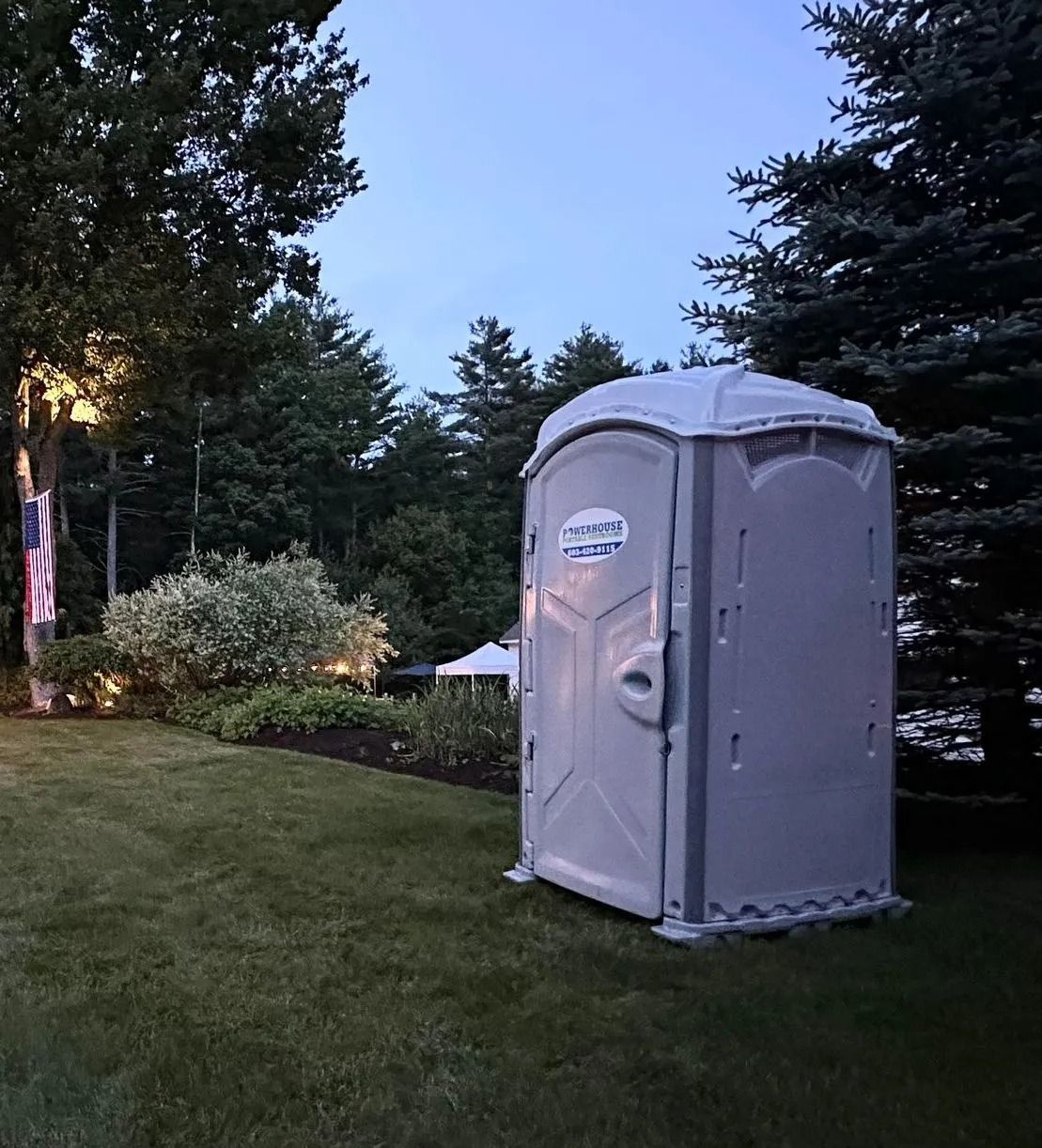 A gray portable restroom sits on a grassy lawn at dusk, with trees and a small white tent in the background.