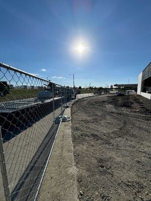 Construction site with chain-link fence, dirt path, and bright sun in a clear blue sky.