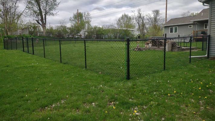 Black chain-link fence surrounding a grassy backyard next to a house under a cloudy sky.