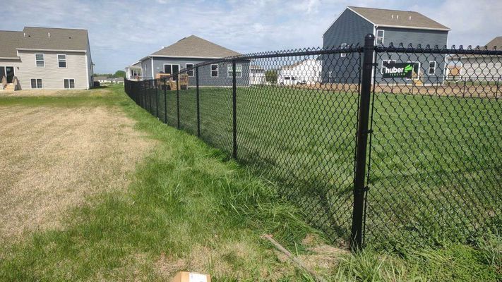 Black chain-link fence between grassy areas, with houses in the background under a blue sky.