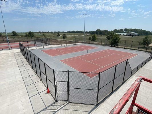 Two red tennis courts surrounded by black fencing, in an outdoor setting.
