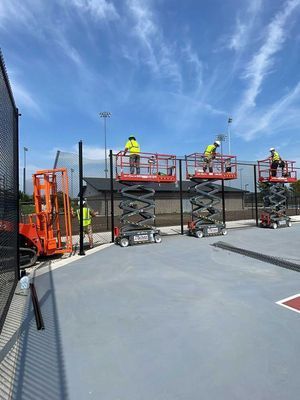 Construction workers installing fencing on a tennis court, using lift platforms. Blue sky overhead.