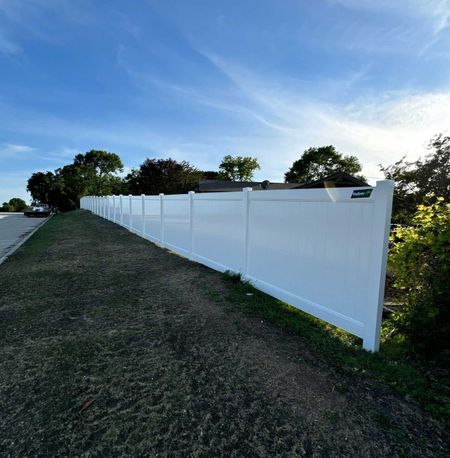 White vinyl fence along grassy area, trees, and blue sky.