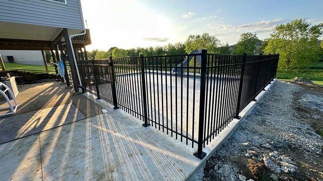 Black metal fence surrounds a pool on a concrete patio, beside a house and a grassy area.
