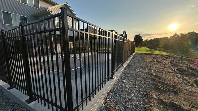 Black metal fence on a retaining wall next to a building with a sunlit field in the background.