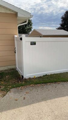 White vinyl fence next to a tan building and a driveway, under a cloudy sky.