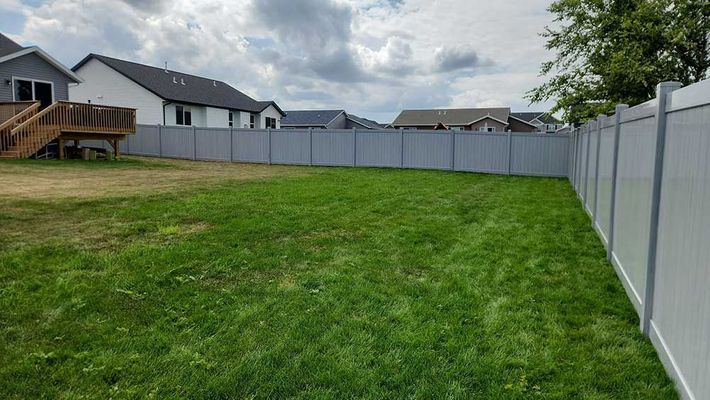 Grassy backyard with a gray privacy fence under a cloudy sky, houses in background.