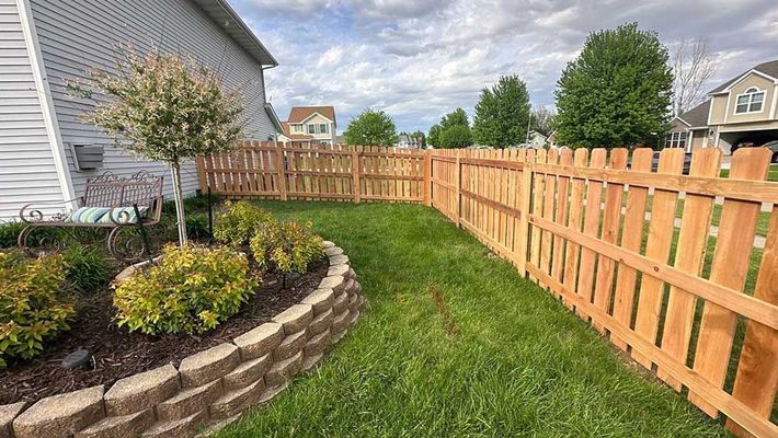 Wooden picket fence enclosing a grassy yard with a small tree and flowerbed. Houses in the background under a cloudy sky.