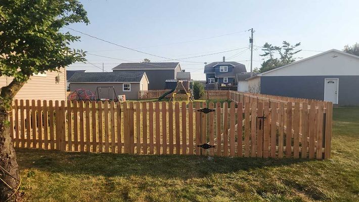 Wooden picket fence encloses a grassy yard with a swing set. Houses and a tree are in the background.
