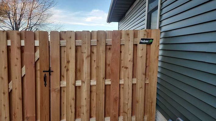 Wooden privacy fence gate next to a blue-sided house on a sunny day.