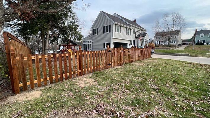 Brown picket fence surrounding a gray house on a grassy lawn; cloudy sky.