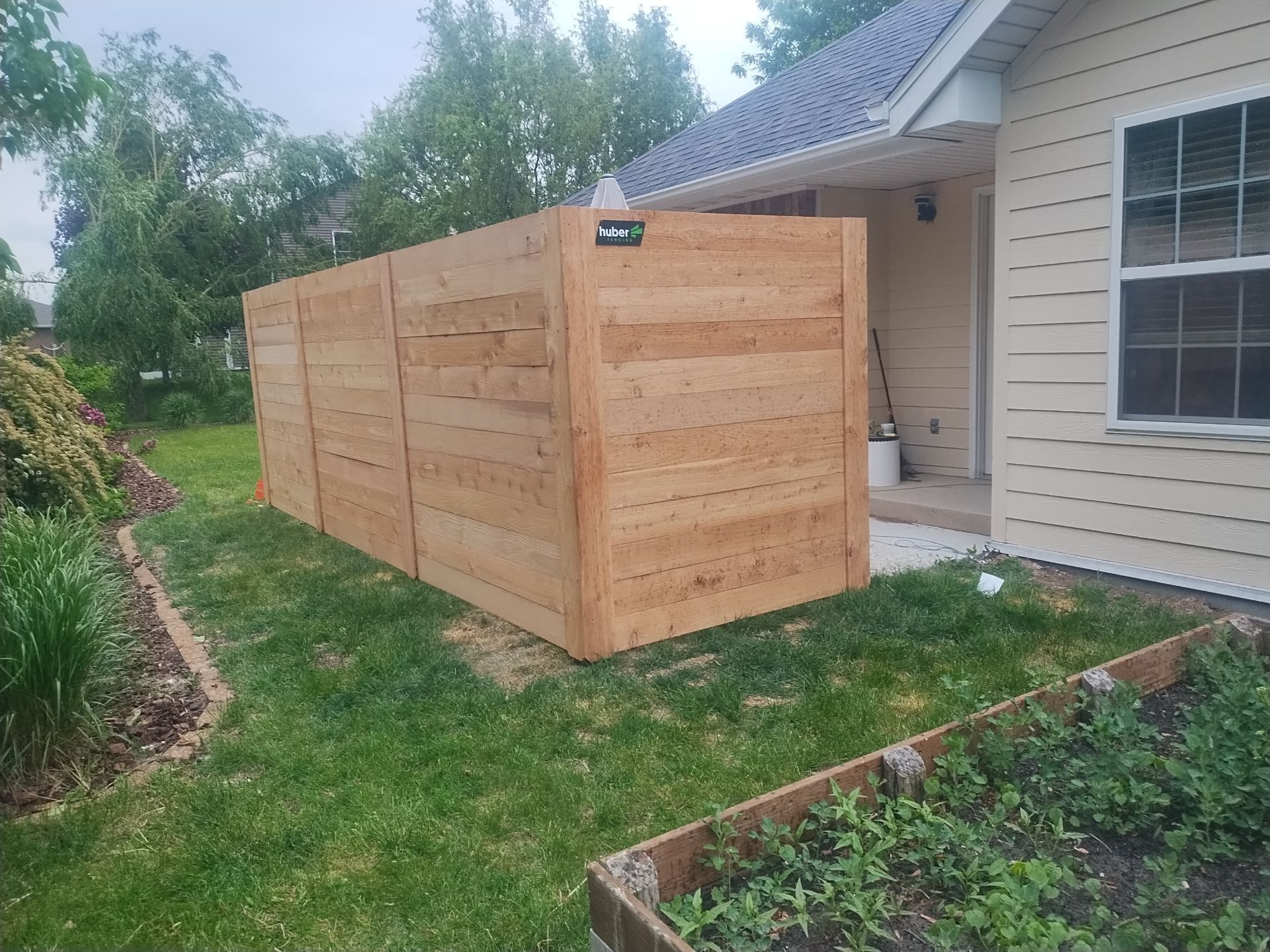 Wooden fence alongside a beige house on green grass. A small garden bed is in the foreground.
