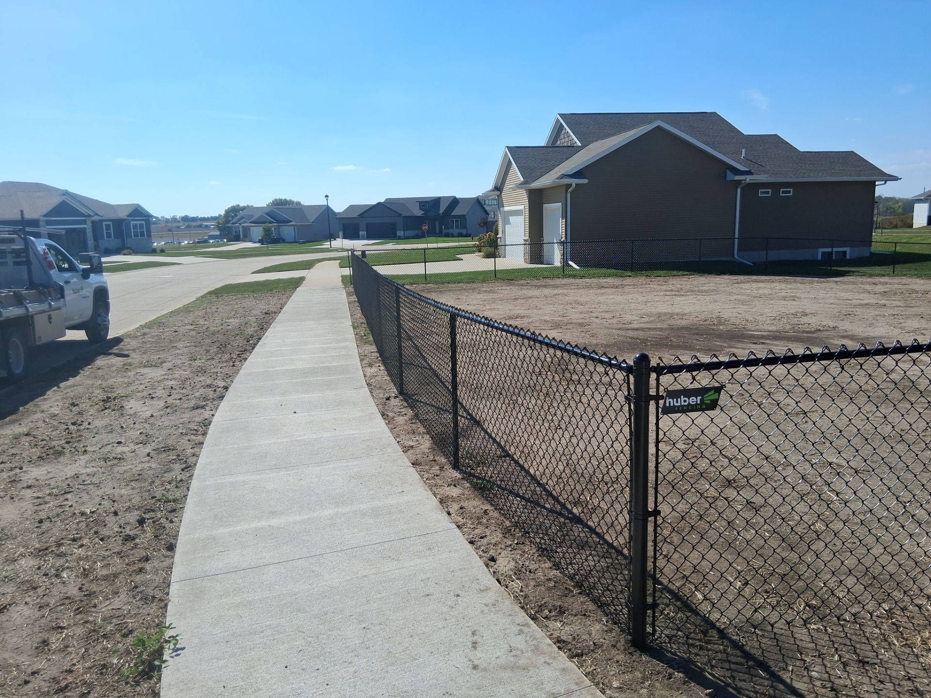 Black chain-link fence bordering a sidewalk next to a construction site, with houses in the background on a sunny day.