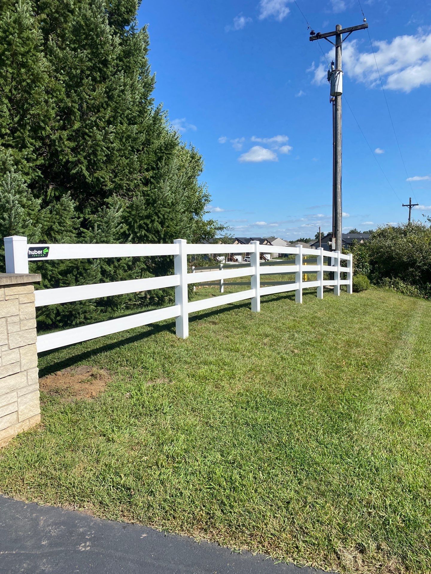White three-rail fence alongside a grassy area under a blue sky, next to a large tree and a utility pole.