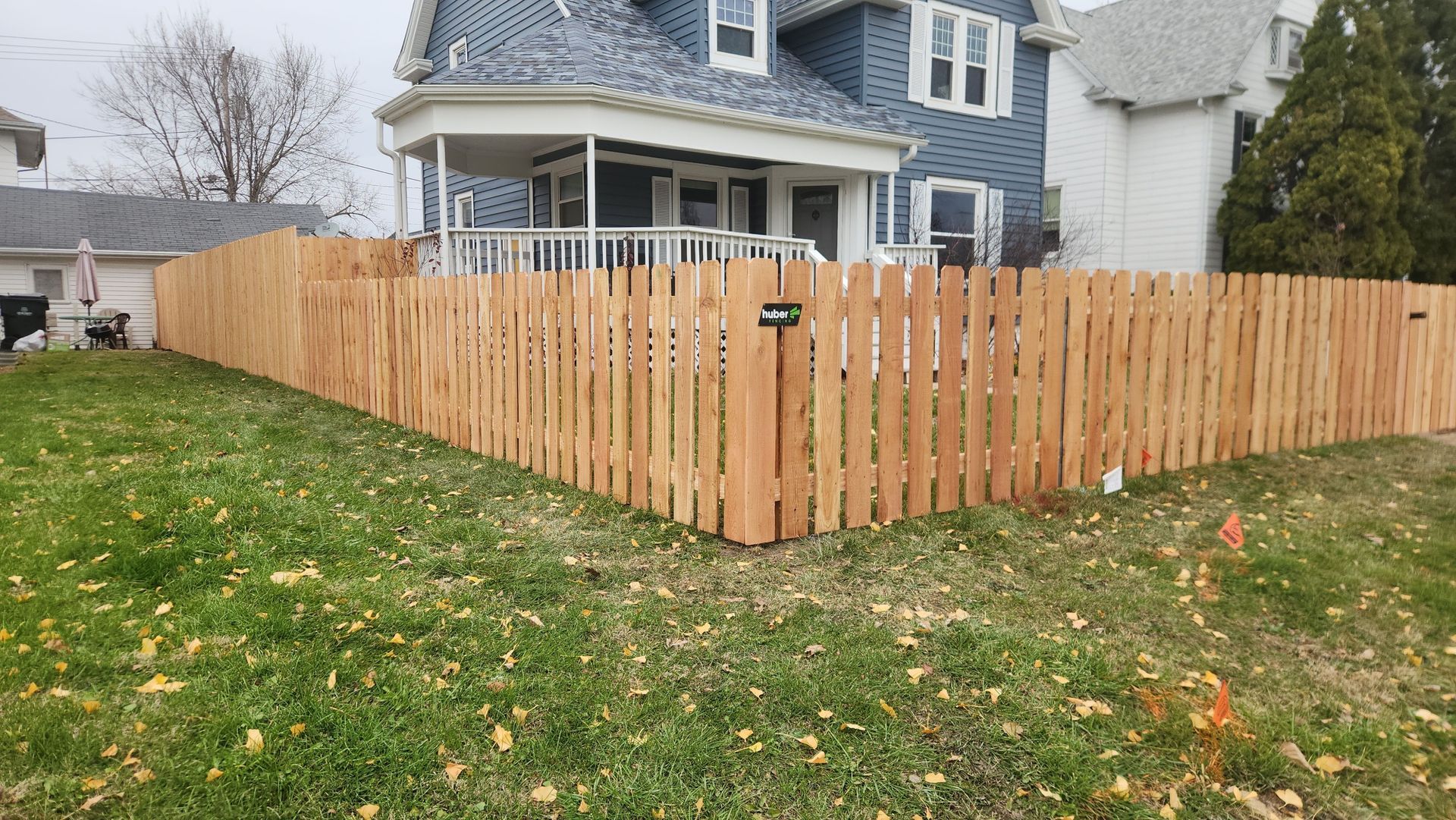 Wooden fence surrounds a house with blue siding. Green grass and scattered leaves are in the foreground.