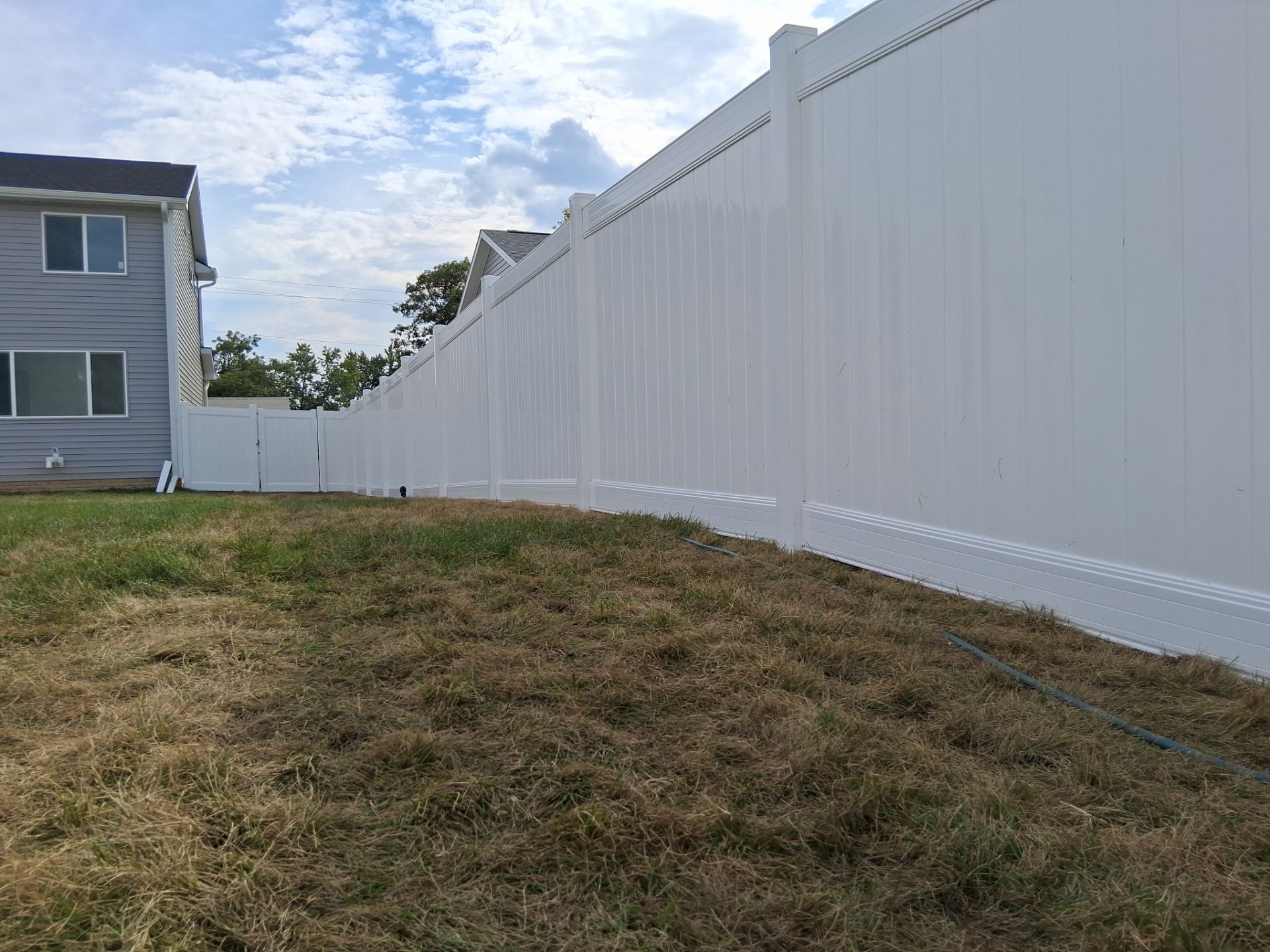 White vinyl fence along dry, grassy yard, with a two-story building in the background against a cloudy sky.