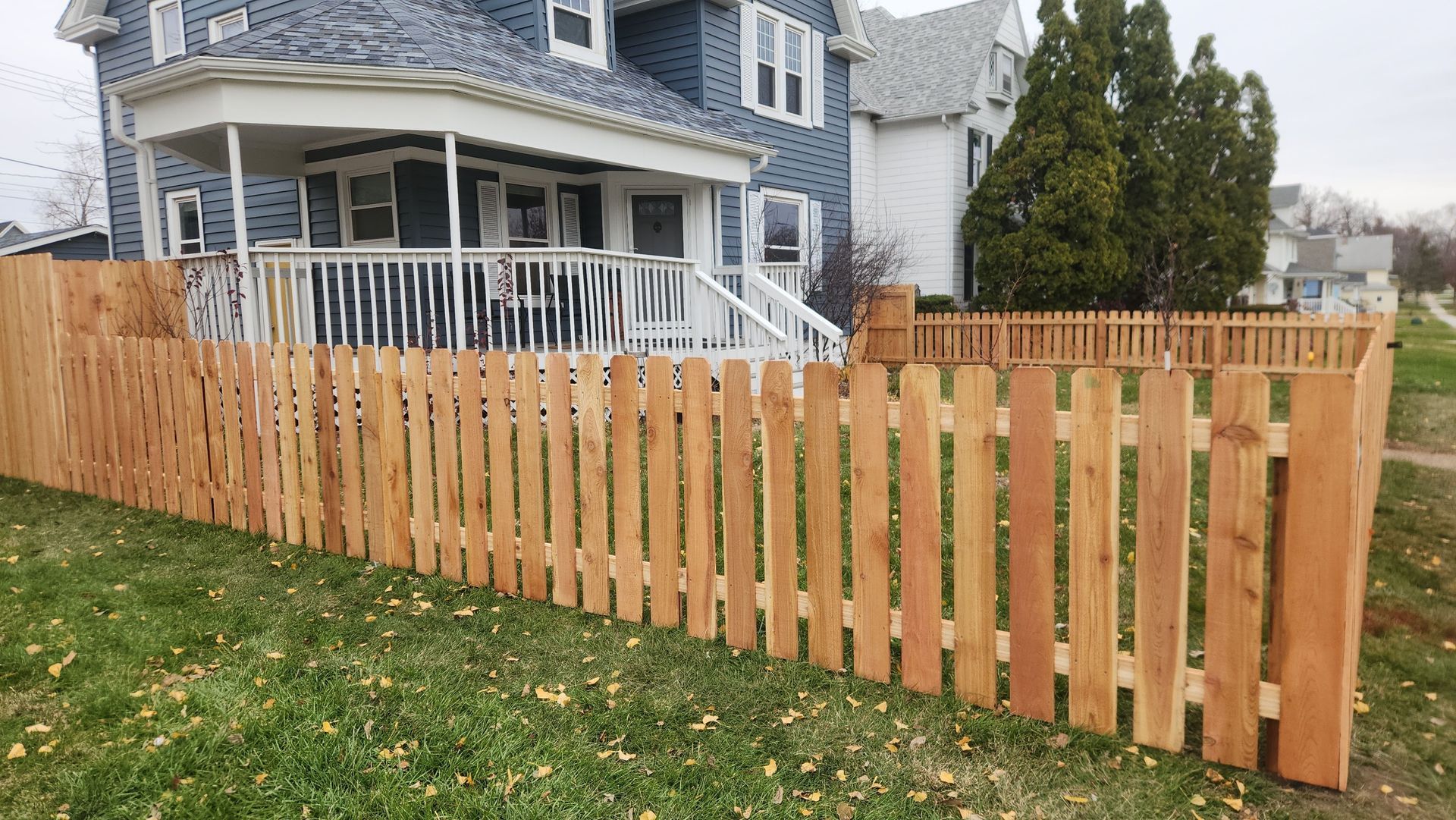Wooden fence in front of a blue house with a white porch. Green grass and overcast sky.