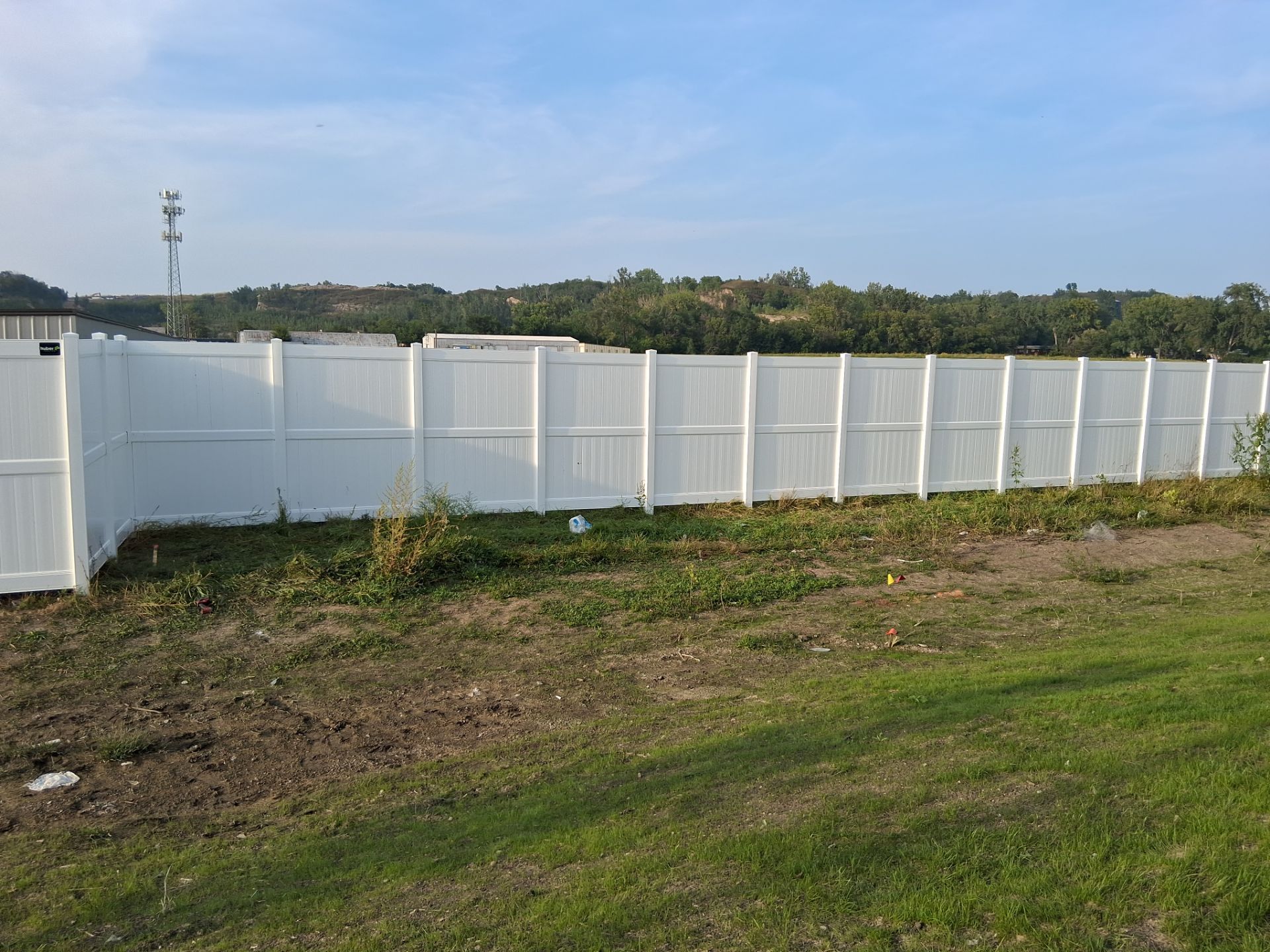 White vinyl fence along grassy area, with trees in the background under a blue sky.