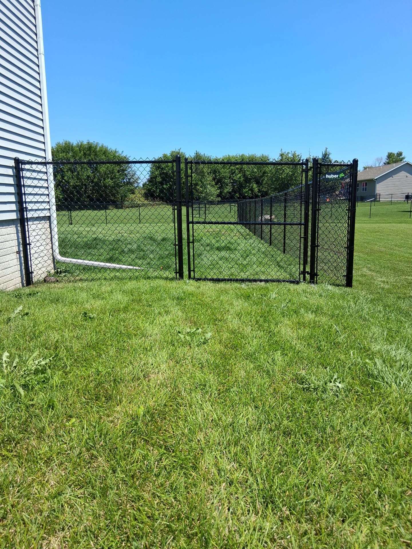 Black chain-link fence with gate, bordering a green lawn near a house. Clear blue sky.