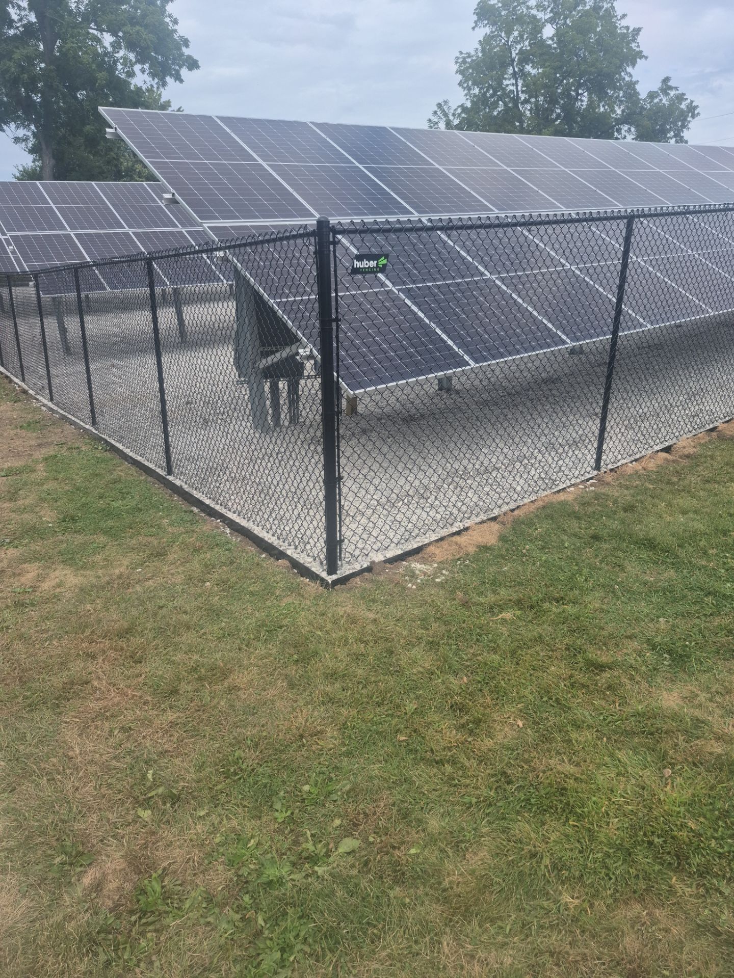 Solar panels surrounded by a black chain-link fence on a gravel bed. Green grass in the foreground, cloudy sky.