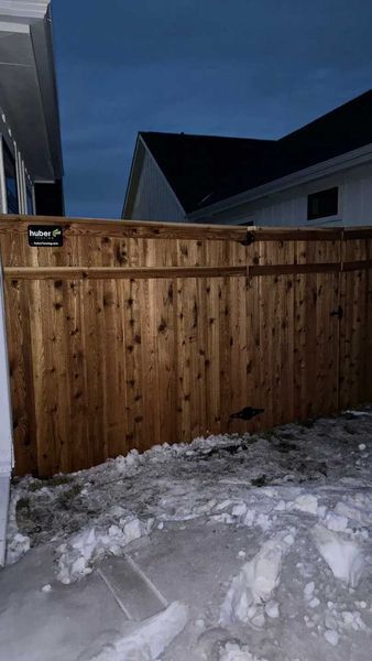 Wooden fence with snow in front, adjacent to a building, under a twilight sky.