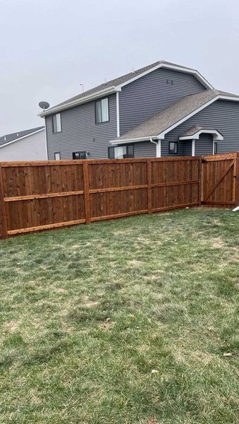 Brown wooden fence surrounds a backyard with a house in the background under a cloudy sky.