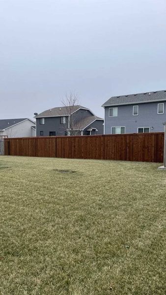 Brown wooden fence in a backyard with green grass and two houses in the background on a cloudy day.