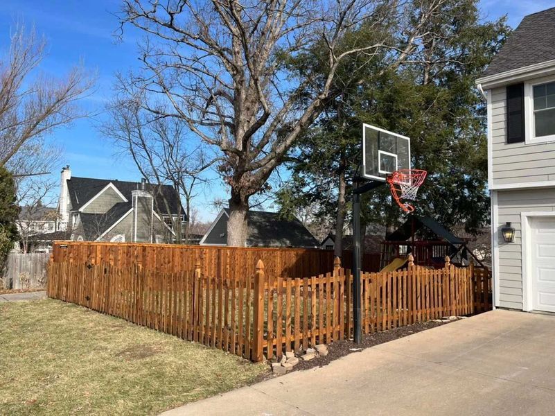 Brown picket fence surrounds a backyard with a basketball hoop and a two-story house.
