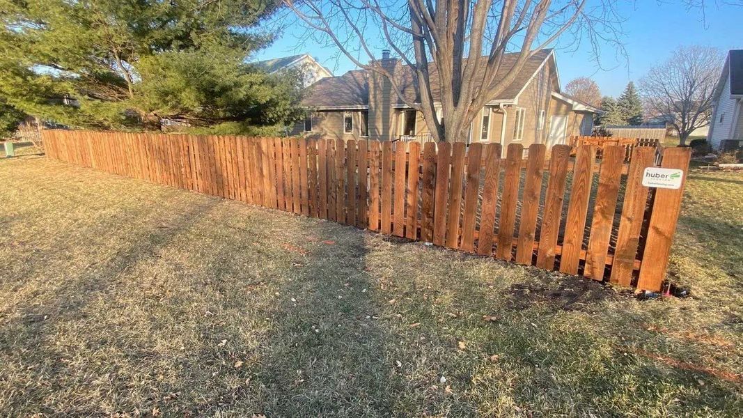 Brown wooden fence in a grassy yard, with a house and trees in the background.