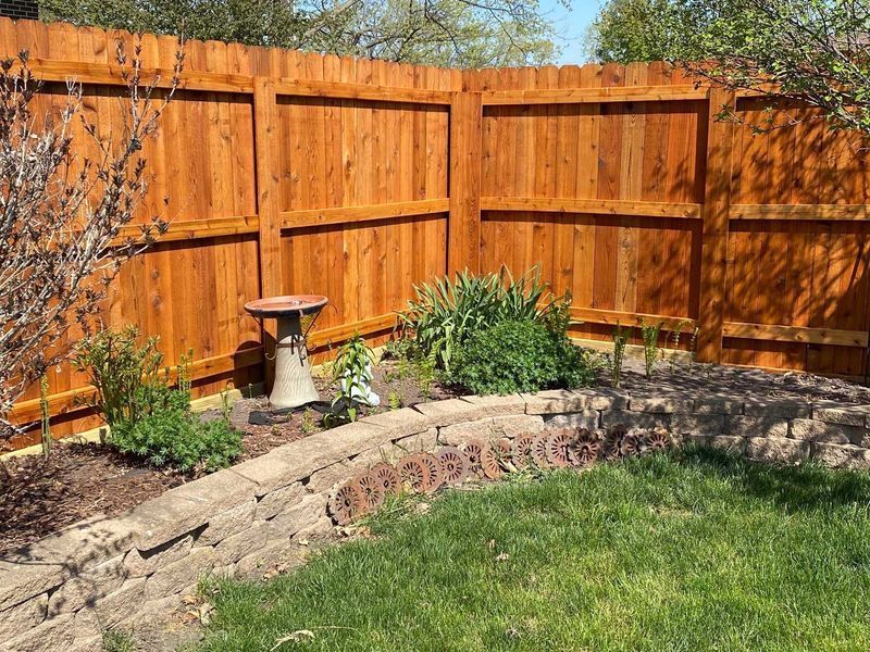 Brown wooden fence enclosing a small garden bed with greenery, a birdbath, and a lawn.