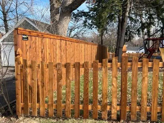 Wooden fence with a contrasting tall privacy fence behind it; trees, sky, and a house in the background.
