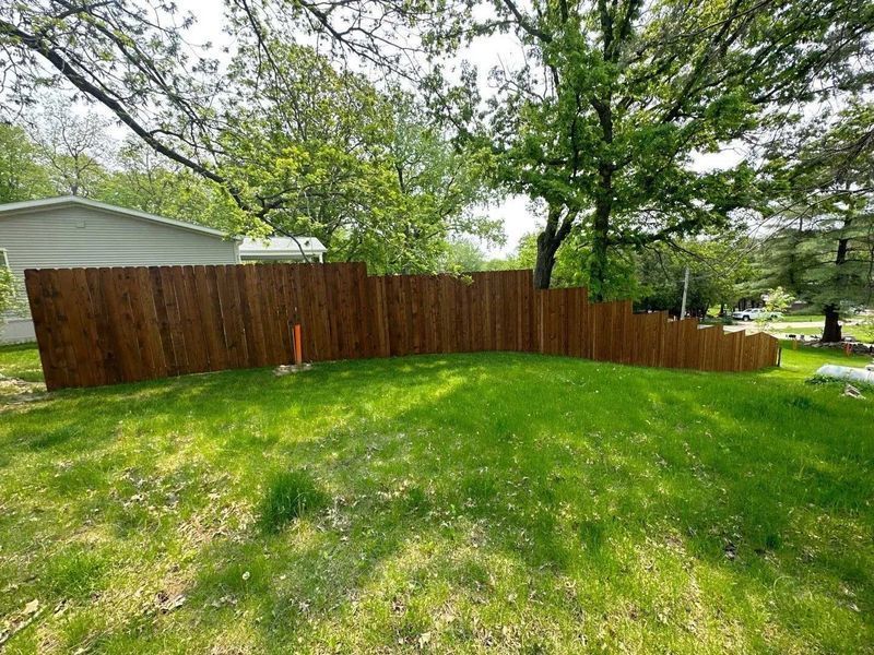 Brown wooden fence curving across a grassy yard; trees and a house in the background.