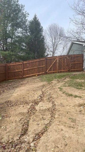 Brown wooden fence with gate in a yard with dirt and bare trees.