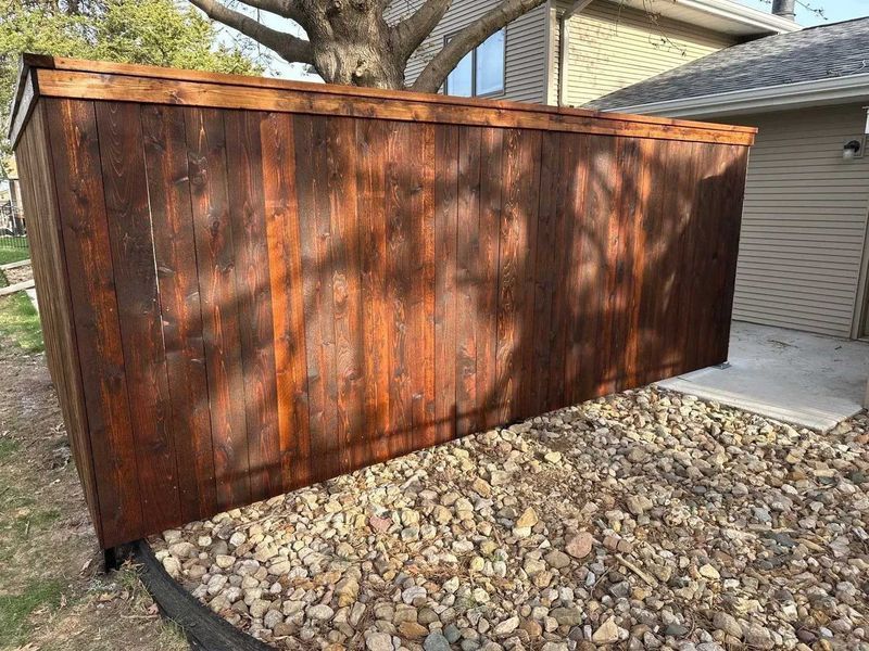 Brown wooden fence with a dark finish, set in a rocky area, near a house and grass.