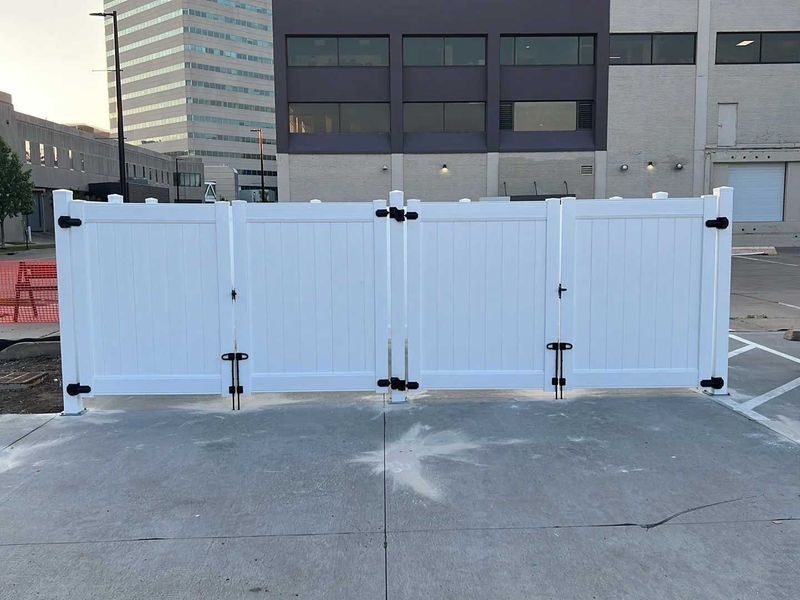 White vinyl fence with four gates in a parking lot; a building is in the background.