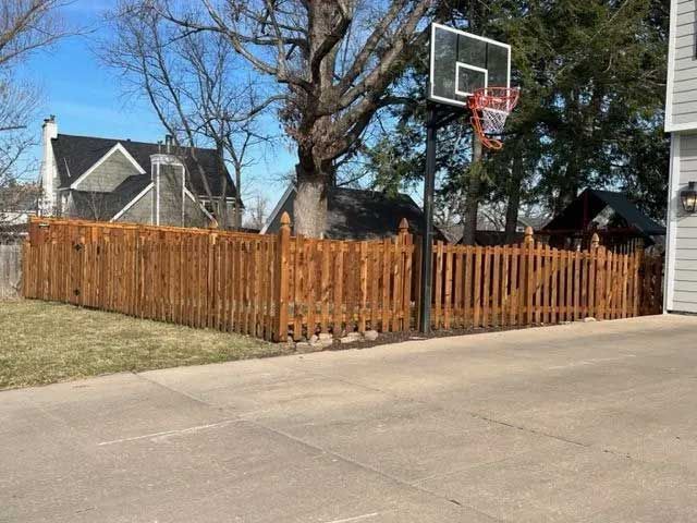 Brown wooden fence surrounds a yard with a basketball hoop, concrete driveway, and houses.