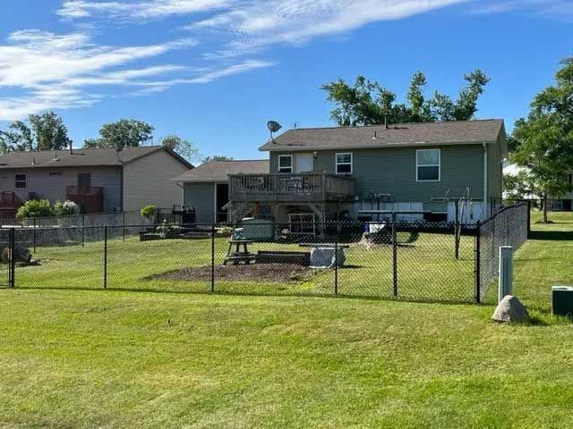 Backyard view of a two-story house with a wooden deck, surrounded by a black fence. Green grass and blue sky.