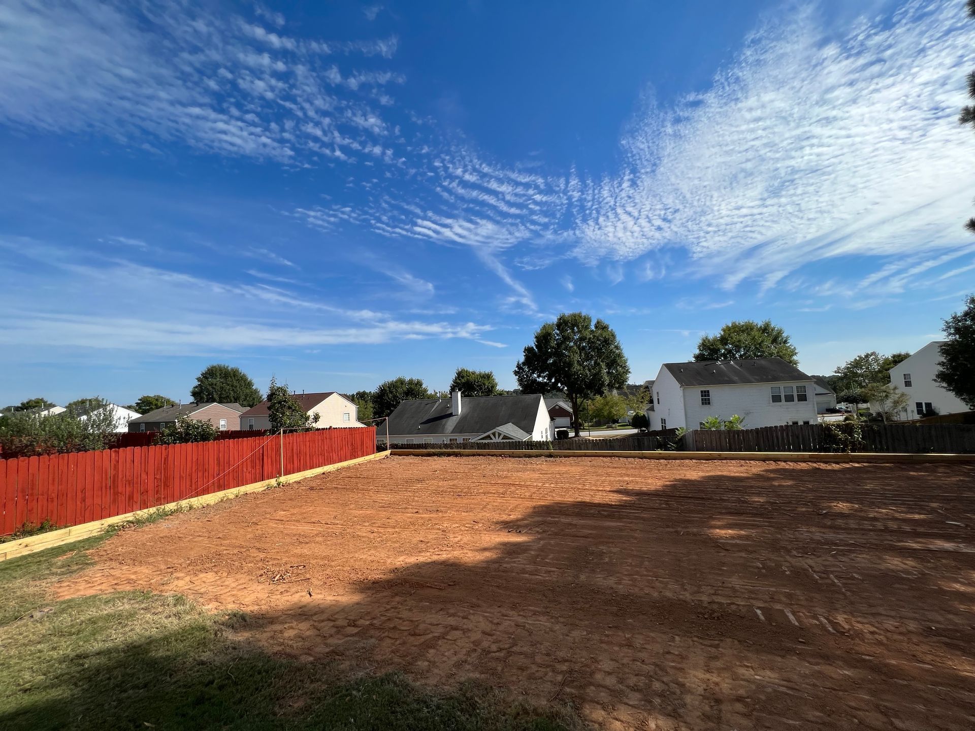 A dirt field with a red fence and houses in the background.