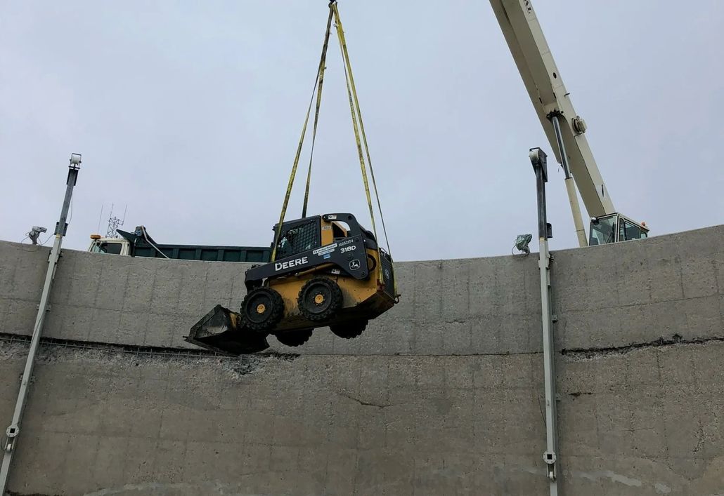 Skid steer loader being lifted by a crane over a concrete wall. Overcast sky.