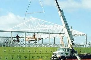 Construction workers using a crane to lift steel beams for a building roof. Bright sky, green siding.
