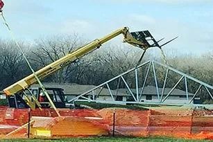 Yellow telehandler placing a metal roof truss on a construction site, with orange fencing and a residential background.