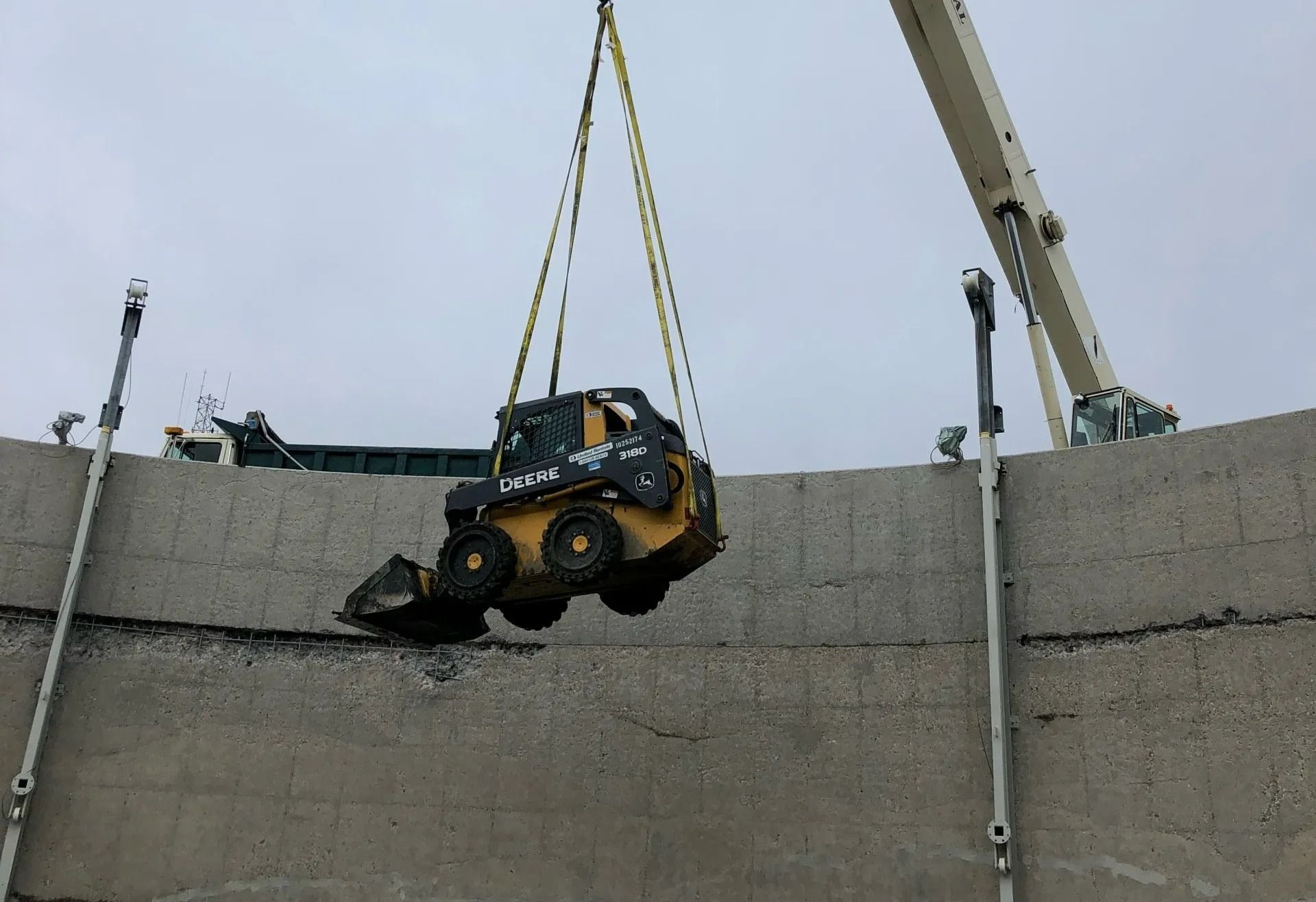 A yellow skid steer being lifted by a crane over a concrete wall.