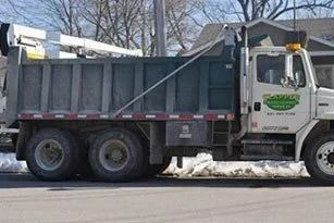Dump truck parked on a street with snow. The truck is white with a green logo, and a raised bed.