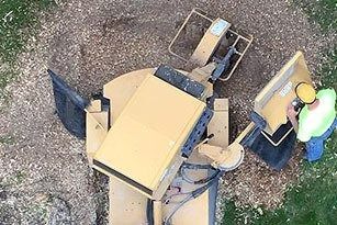 Overhead view of a yellow stump grinder and a worker in a hard hat and safety vest inspecting it.